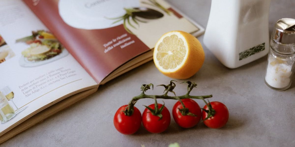 Flat lay of cooking ingredients including tomatoes and a lemon with a cookbook.